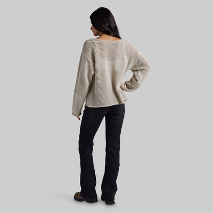 A woman shown from the back wearing a cream-colored, textured knit long-sleeve sweater and black pants, standing against a solid light gray background.