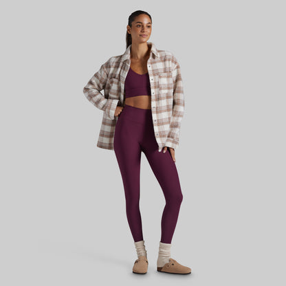 A woman modeling a matching athletic set consisting of a deep purple sports bra and high-waisted leggings, styled with an open beige and brown plaid flannel shirt, cream knit socks, and tan slip-on shoes against a solid gray background.