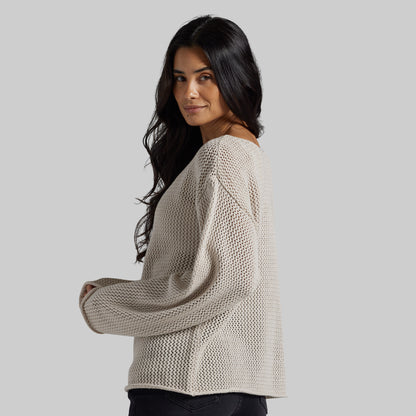 A woman wearing a beige knit sweater with a textured open-knit pattern, shown from a three-quarter back view against a solid gray background.