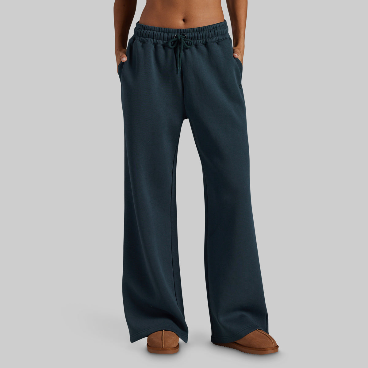 A person wearing dark teal wide-leg sweatpants with an elastic drawstring waistband and side pockets, paired with brown slippers, shown against a plain light gray studio background.