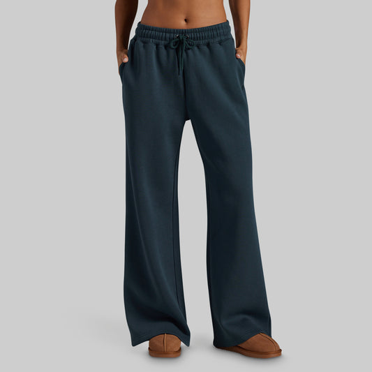 A person wearing dark teal wide-leg sweatpants with an elastic drawstring waistband and side pockets, paired with brown slippers, shown against a plain light gray studio background.