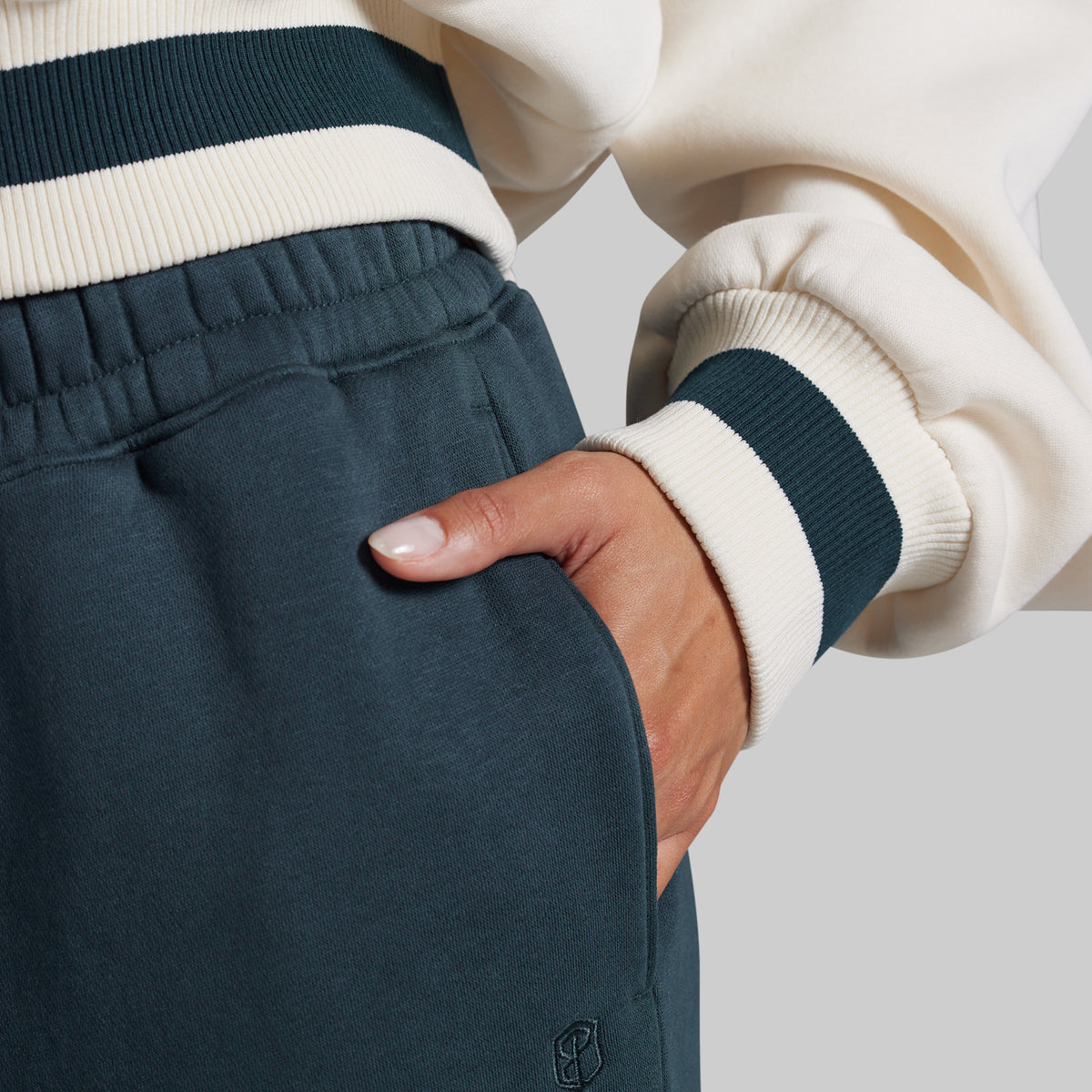 Close-up of a person wearing dark teal fleece sweatpants with a side pocket and a cream and dark teal ribbed sweatshirt, with the hand in the pocket.