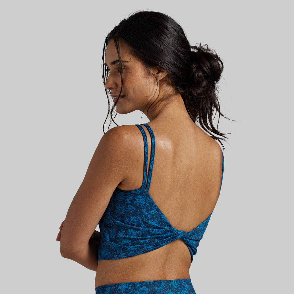 A woman shown from the back wearing a blue patterned sports bra with double shoulder straps and a twisted back detail, against a solid gray background.