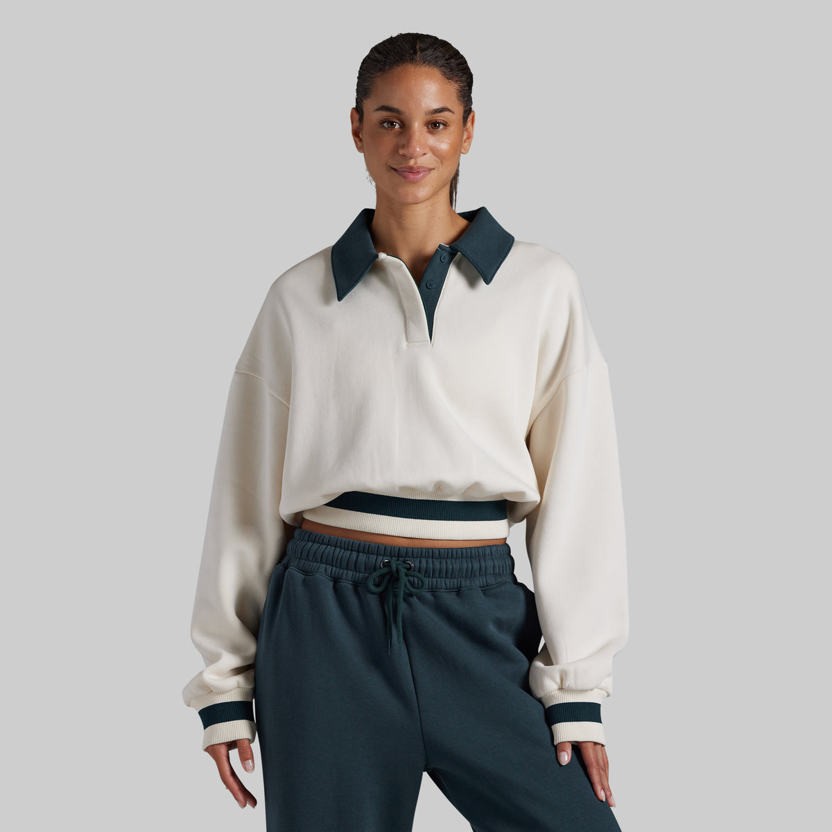 A woman modeling a cropped long-sleeve polo-style top in off-white with dark teal contrast collar, placket, and cuffs, paired with dark teal drawstring sweatpants, against a solid light gray studio background.