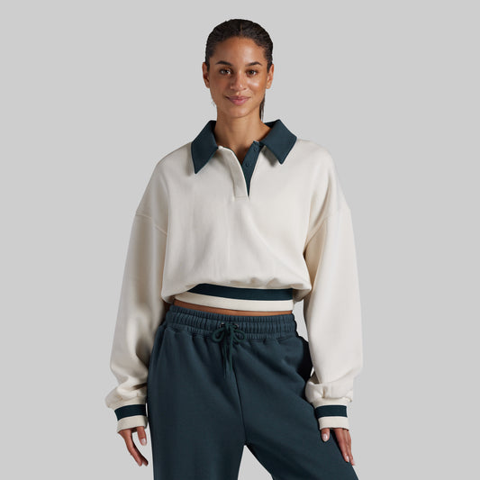 A woman modeling a cropped long-sleeve polo-style top in off-white with dark teal contrast collar, placket, and cuffs, paired with dark teal drawstring sweatpants, against a solid light gray studio background.