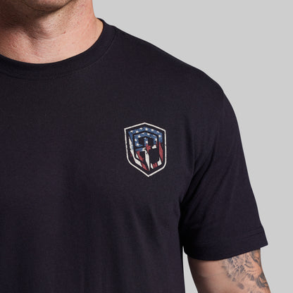 Black t-shirt with a shield emblem featuring American flag colors on a gray background