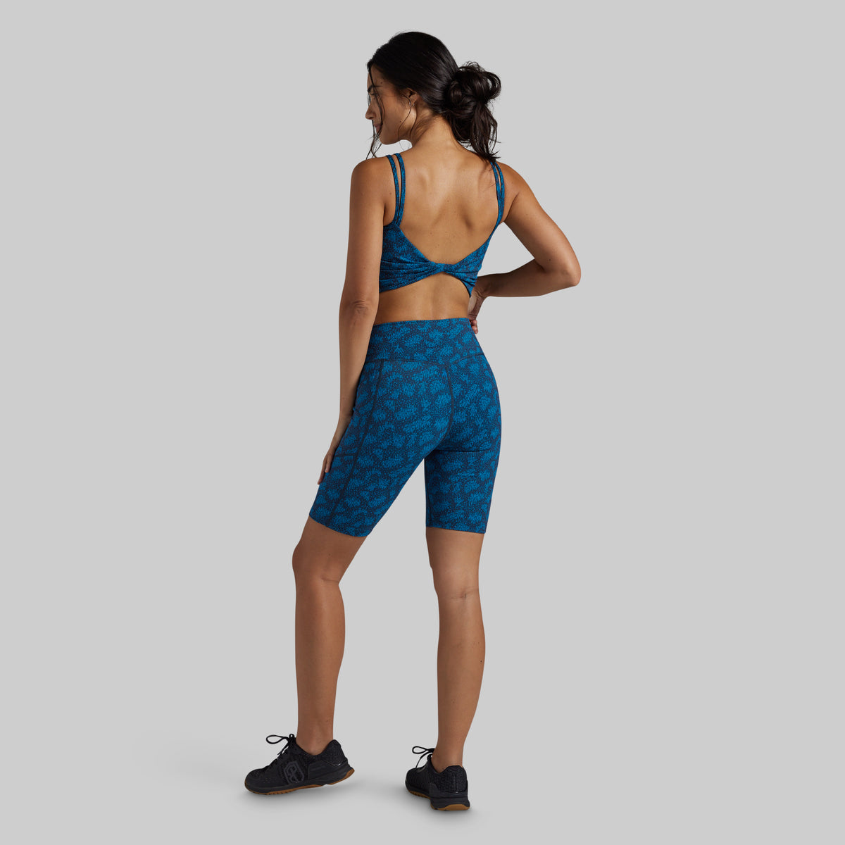 A woman modeling a matching blue patterned activewear set, featuring a sports bra and high-waisted biker shorts, shown from a rear three-quarter view against a solid gray background.