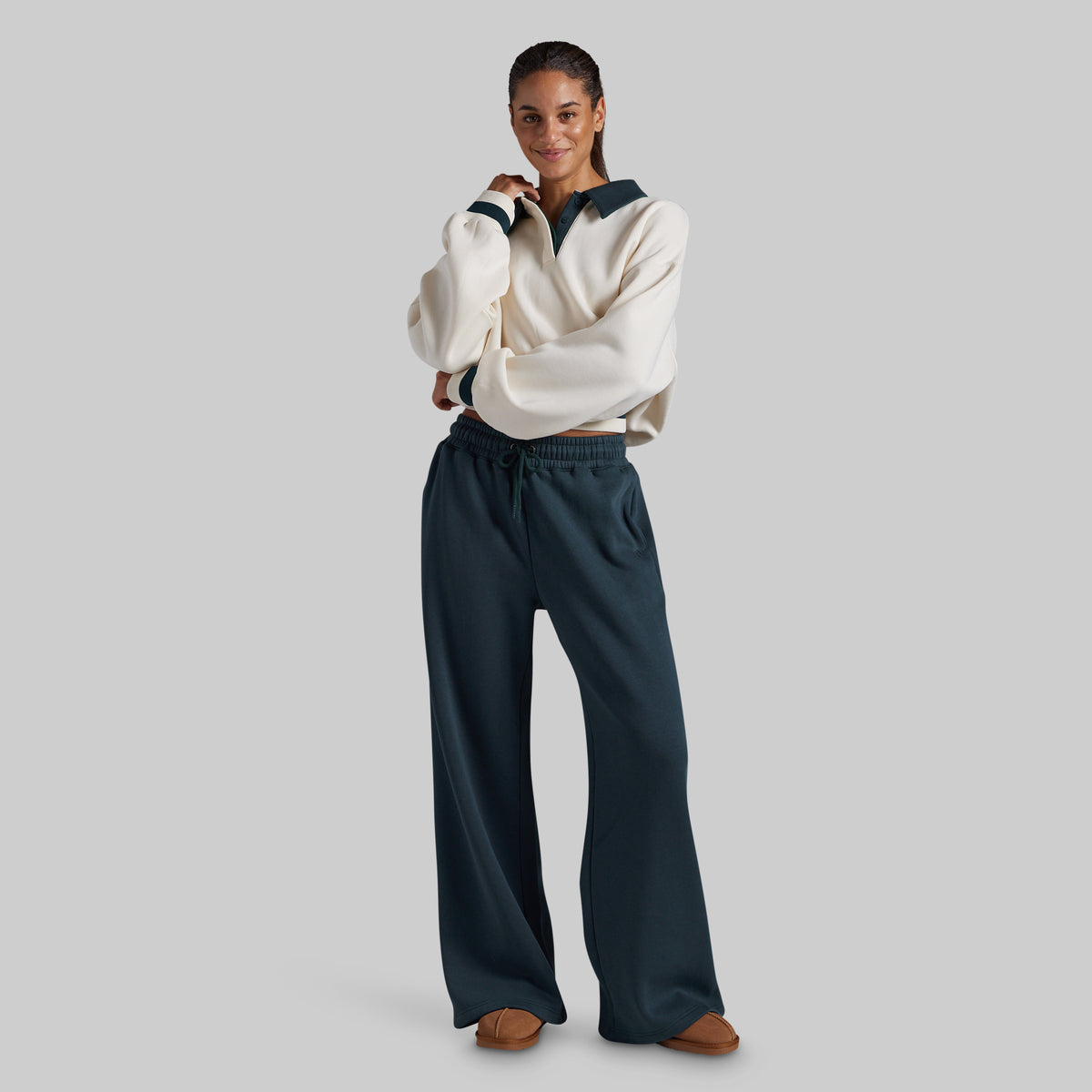 A woman modeling a cream sweatshirt with dark teal trim and matching dark teal wide-leg sweatpants, standing against a solid light gray studio background.