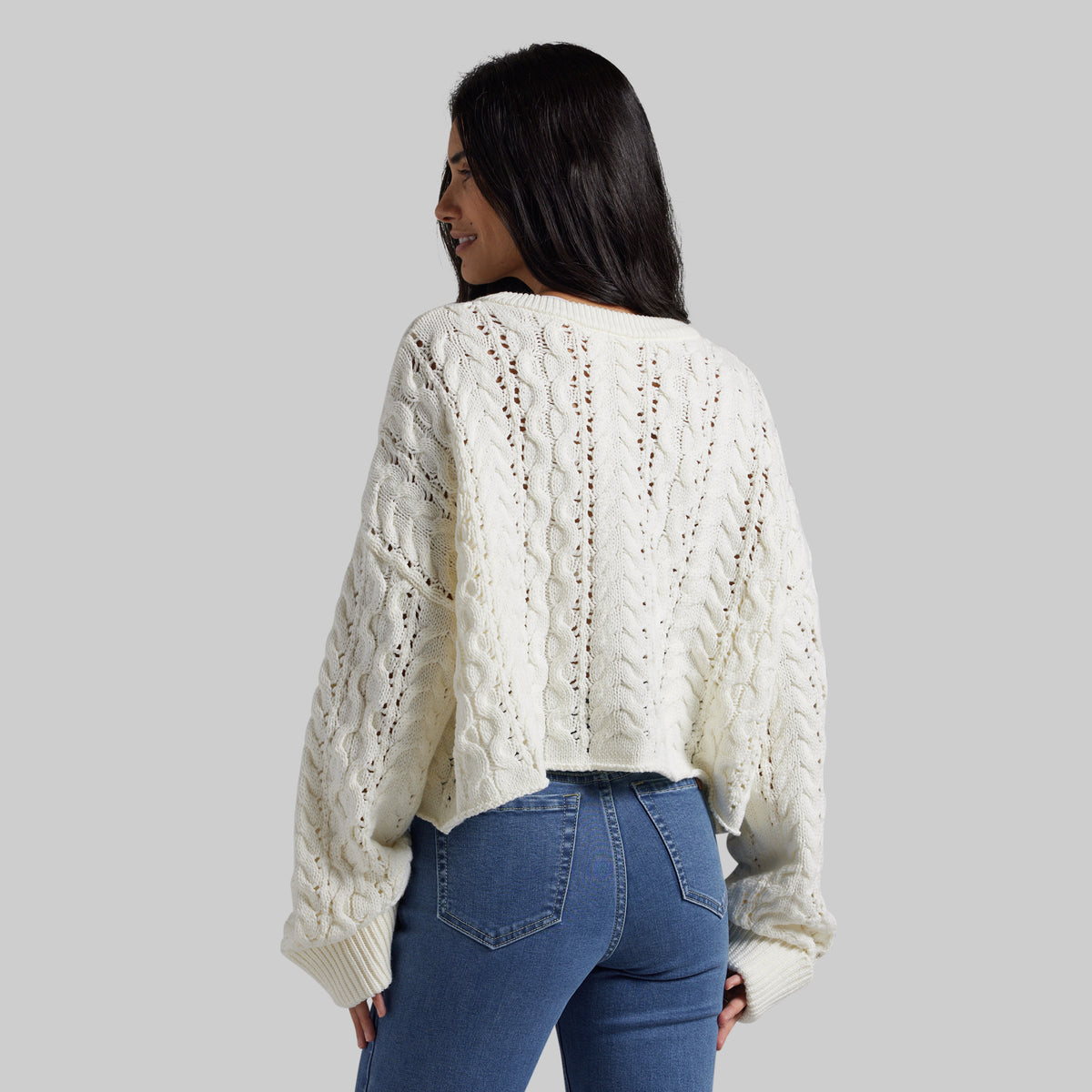 A woman shown from the back wearing a cream cable-knit cropped sweater and blue jeans against a solid gray studio background.