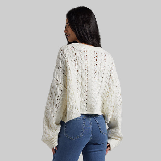 A woman shown from the back wearing a cream cable-knit cropped sweater and blue jeans against a solid gray studio background.