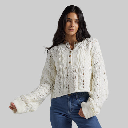 A woman wearing a cream cable-knit cropped sweater with a buttoned collar and blue jeans, photographed against a solid light gray studio background.