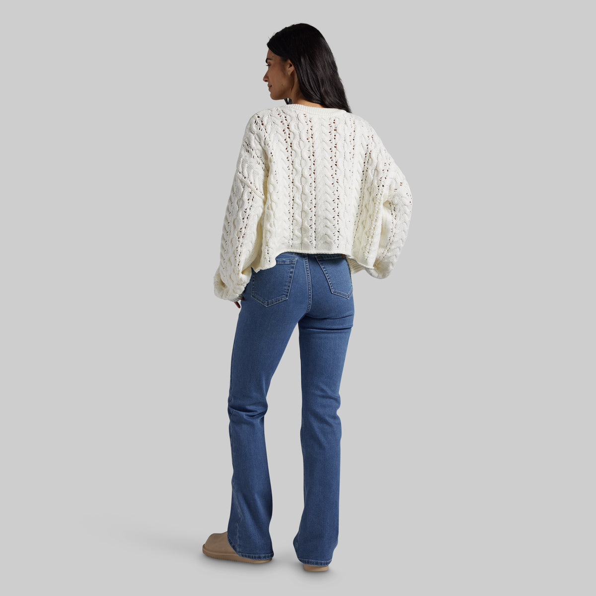 A woman shown from the back wearing a cream cable-knit cropped sweater and blue jeans, standing against a solid gray studio background.