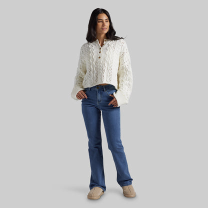 A woman modeling a cream cable-knit cropped sweater with blue jeans and tan shoes, standing against a solid gray studio background.