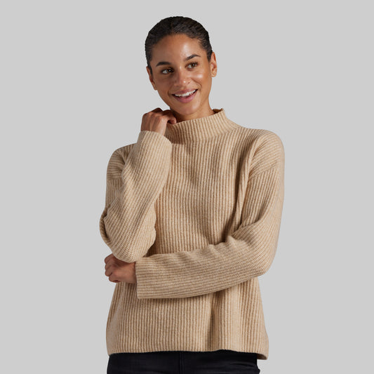 A woman wearing a beige ribbed knit mock neck sweater, posing against a solid gray studio background.