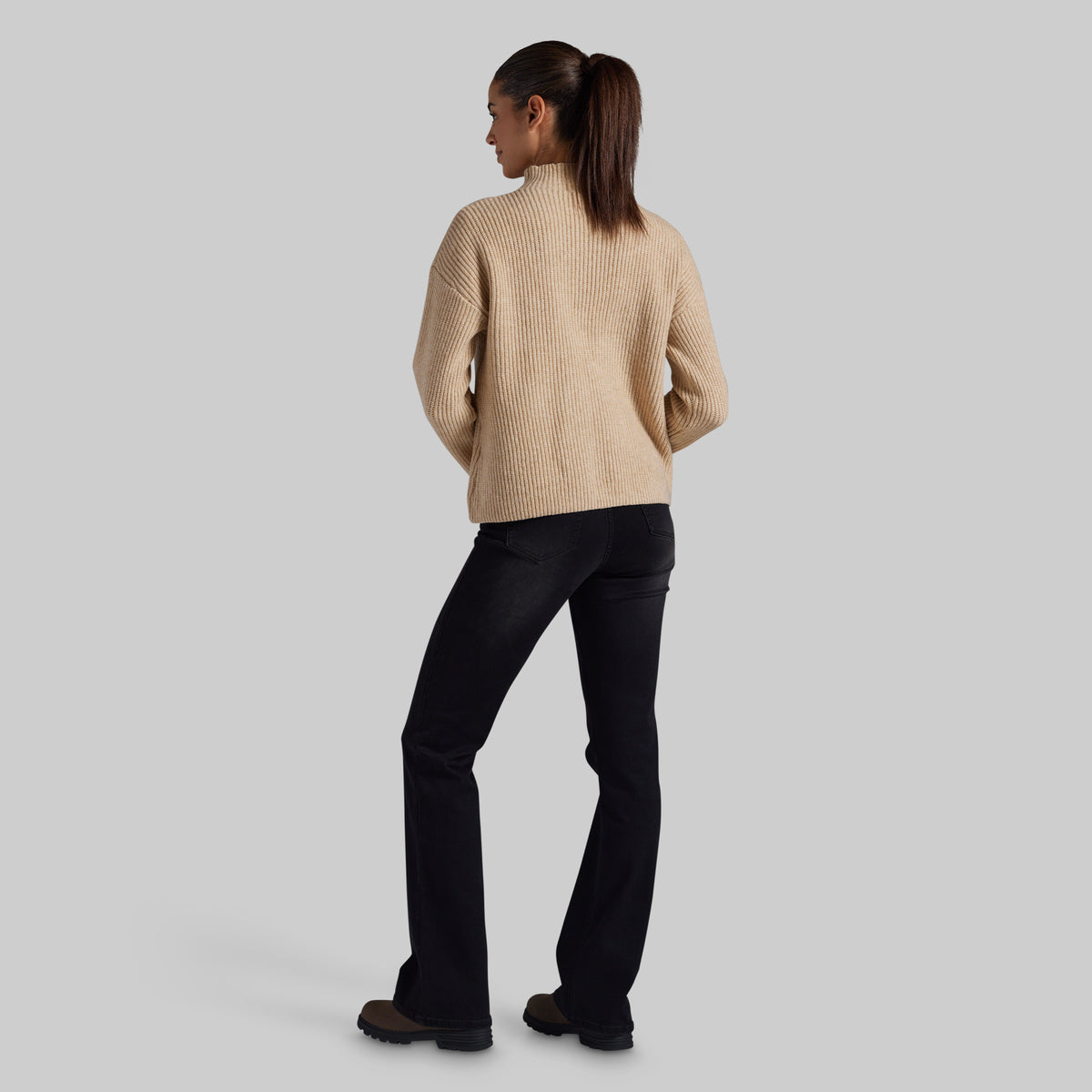 A woman shown from the back wearing a beige ribbed knit mock-neck sweater and black jeans, standing against a solid light gray studio background.