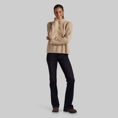 A woman modeling a beige ribbed knit mock-neck sweater and black jeans against a solid light gray background.