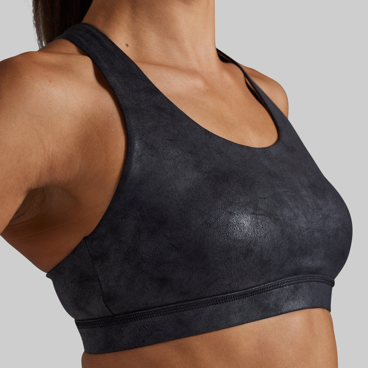 Close-up of a woman wearing a dark charcoal gray sports bra with a textured, mottled finish against a solid light gray background.