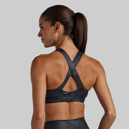 A woman shown from the back wearing a dark charcoal gray sports bra with a crisscross crossover strap design on a solid light gray background.