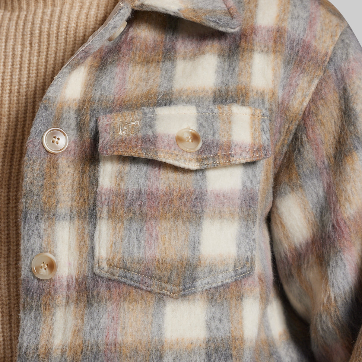 Close-up of a fuzzy plaid jacket with a chest pocket, featuring a cream, gray, and muted pink pattern, worn over a tan ribbed knit sweater.