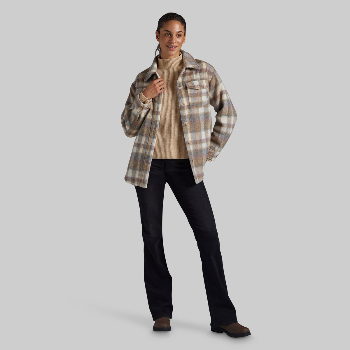 A woman modeling a beige and brown plaid button-up shirt jacket over a beige ribbed turtleneck sweater, paired with black pants and brown shoes, against a solid gray background.