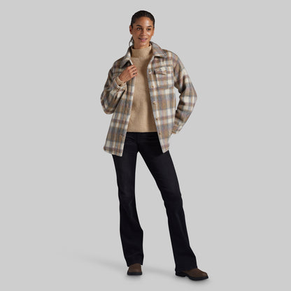 A woman modeling a beige and brown plaid button-up shirt jacket over a beige ribbed turtleneck sweater, paired with black pants and brown shoes, against a solid gray background.
