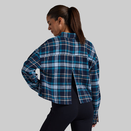 A woman shown from the back wearing a cropped, long-sleeve plaid button-up shirt in navy, teal, and light blue, paired with black leggings, against a solid gray studio background.