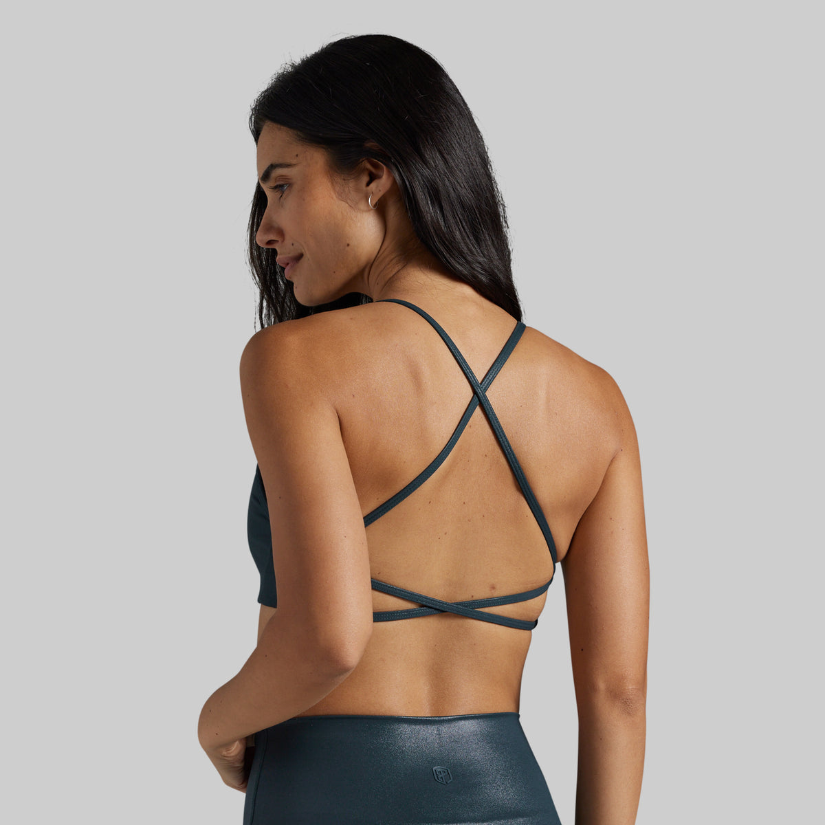 A woman shown from the back wearing a dark teal sports bra with crisscrossing thin straps and matching high-waisted leggings against a solid gray background.