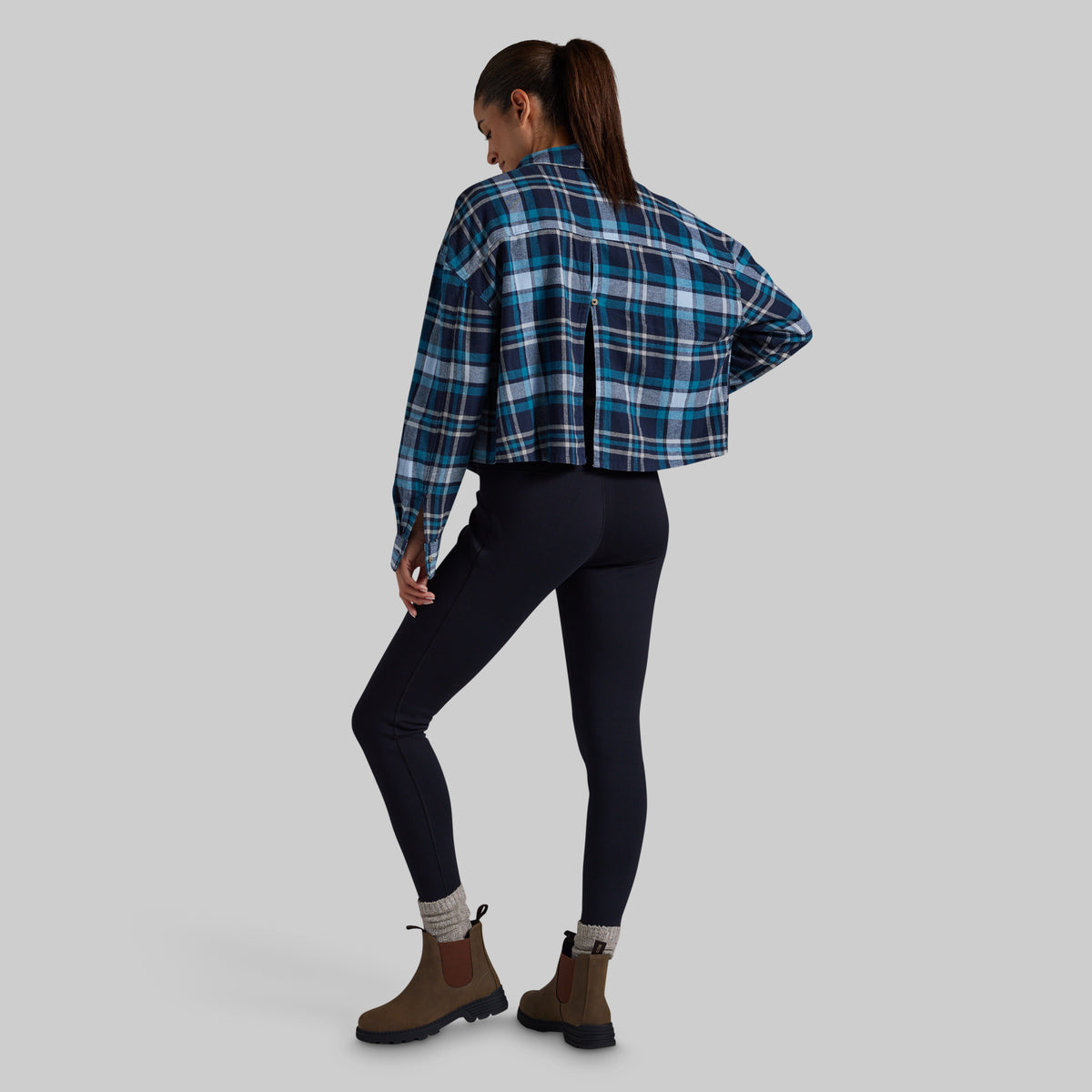 A woman is shown from the back wearing a cropped blue and navy plaid flannel shirt, dark navy leggings, and brown Chelsea boots against a solid gray studio background.