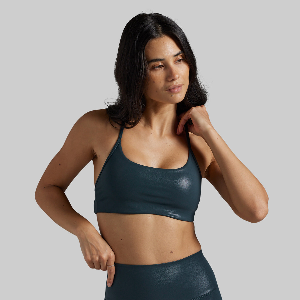 A woman modeling a dark teal metallic sports bra and matching high-waisted leggings against a solid gray studio background.