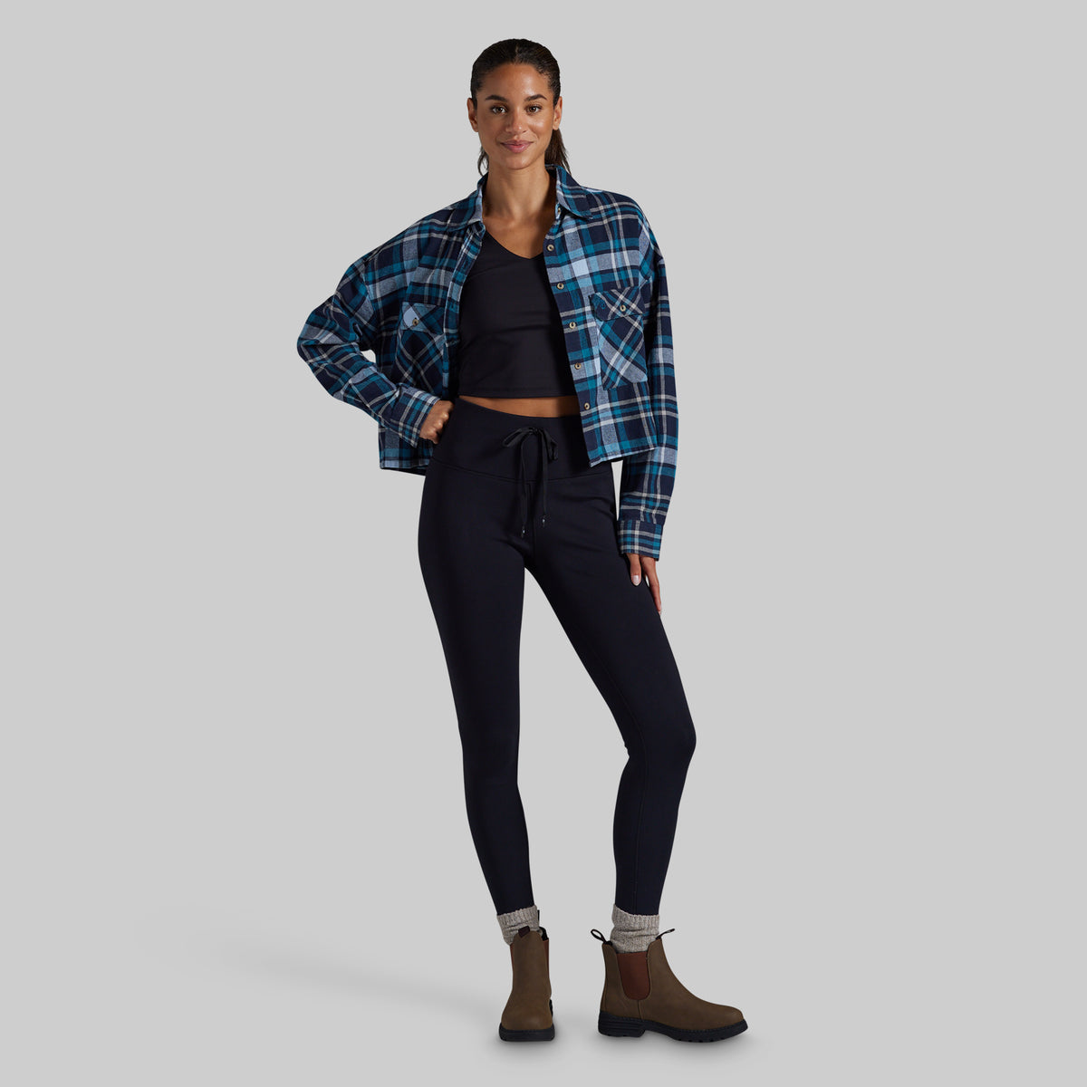 A model stands against a solid gray background wearing a blue and black plaid flannel shirt over a black crop top, black pants, and brown Chelsea boots.