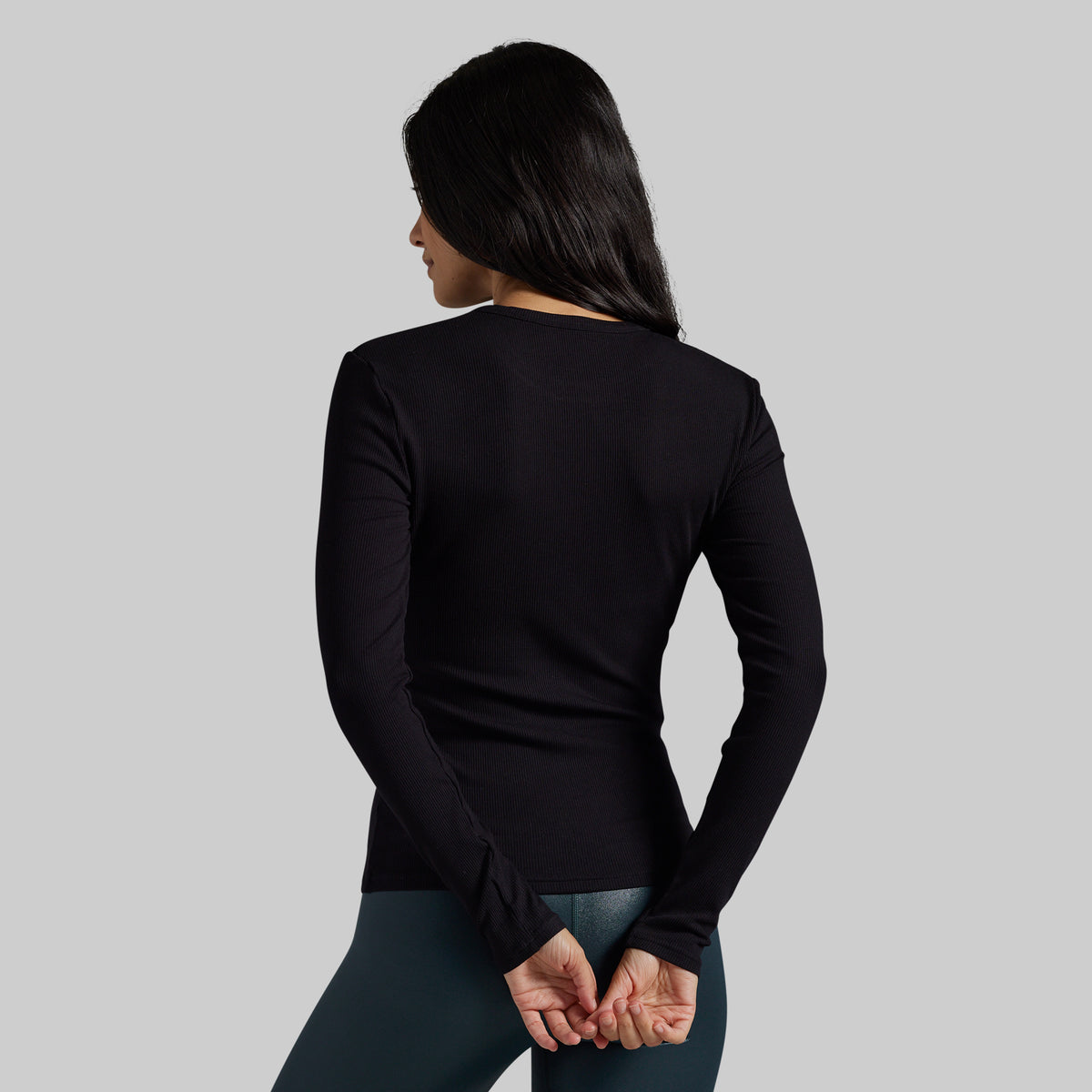 A woman shown from the back wearing a fitted black long-sleeve top and dark leggings against a plain light gray studio background.