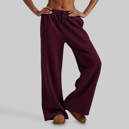 A model wearing wide-leg lounge pants in deep burgundy, paired with brown suede slippers, against a solid gray studio background.