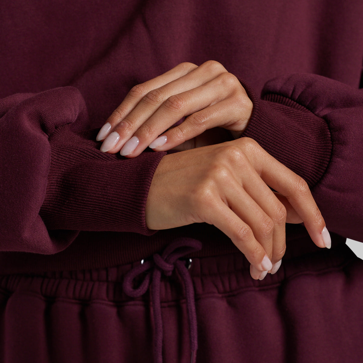 Close-up of a person wearing a deep burgundy sweatshirt and matching sweatpants, with both hands crossed over the chest. The image highlights the ribbed knit cuffs and the drawstring waistband.