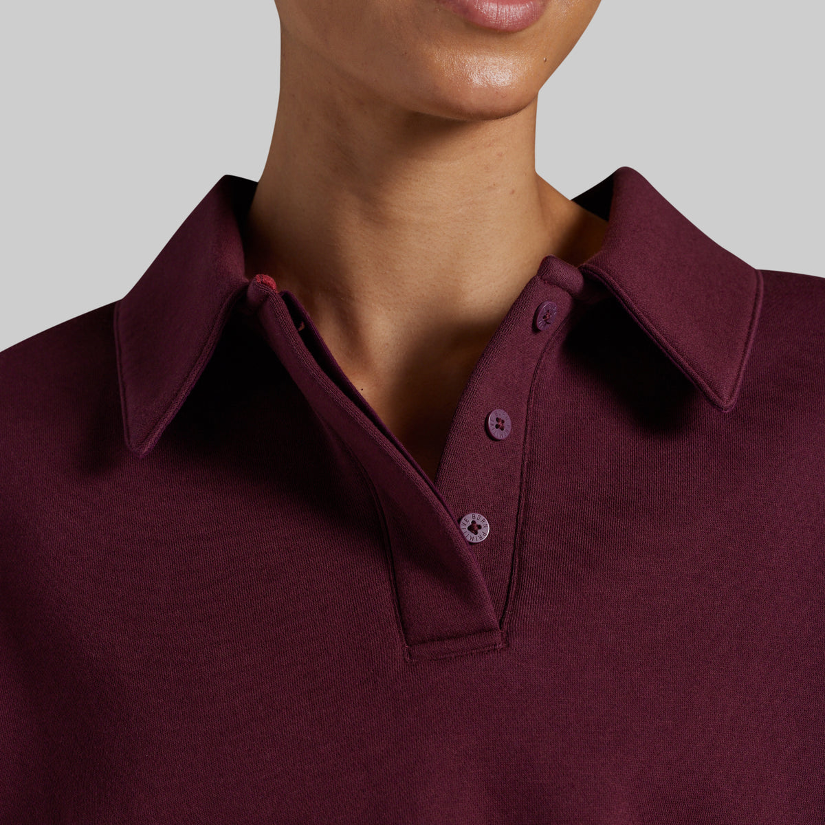 Close-up of a person wearing a deep burgundy polo sweatshirt with a classic collar and matching buttons, shown against a neutral gray background.