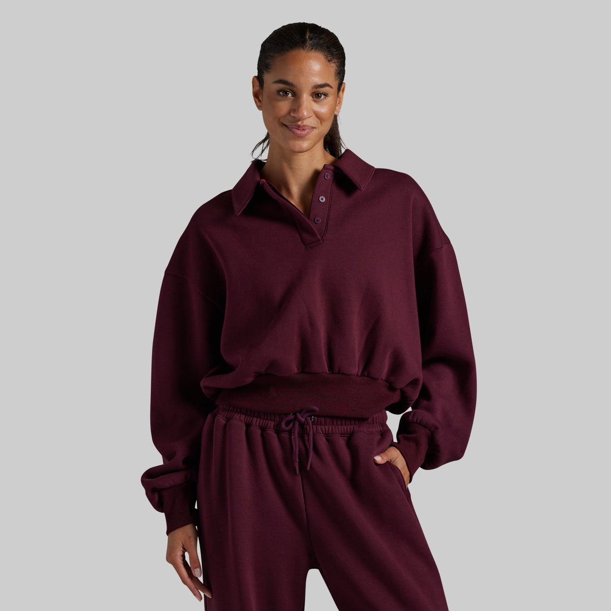 A woman modeling a matching burgundy lounge set consisting of a long-sleeve collared sweatshirt with a button placket and matching drawstring sweatpants, set against a solid gray background.