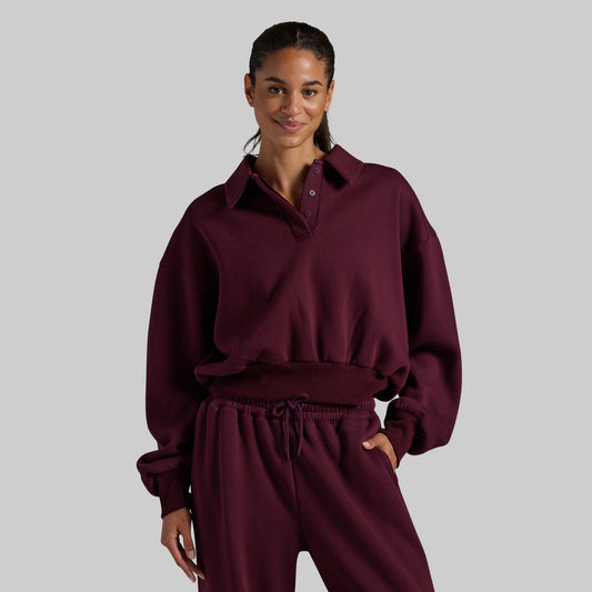 A woman modeling a matching burgundy lounge set consisting of a long-sleeve collared sweatshirt with a button placket and matching drawstring sweatpants, set against a solid gray background.