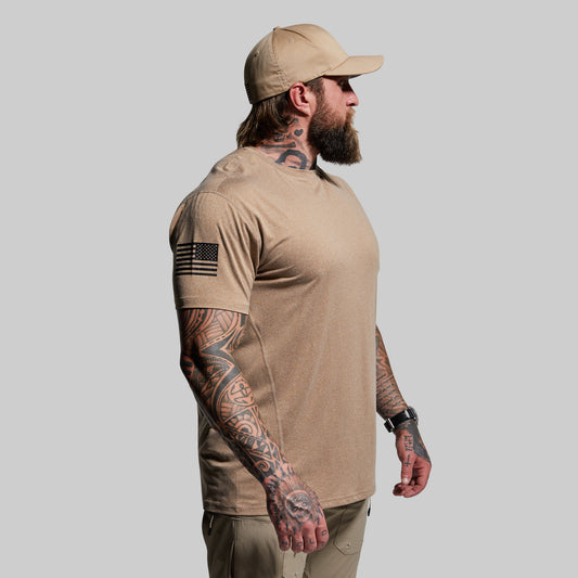 Man wearing a beige t-shirt and cap with tattoos on arms, standing against a gray background