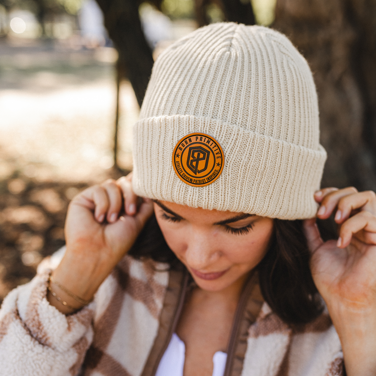 A woman outdoors wearing a cream ribbed knit beanie with a circular tan leather patch on the front. She is adjusting the beanie with both hands, wearing a brown and cream plaid fleece jacket, with a blurred park background.