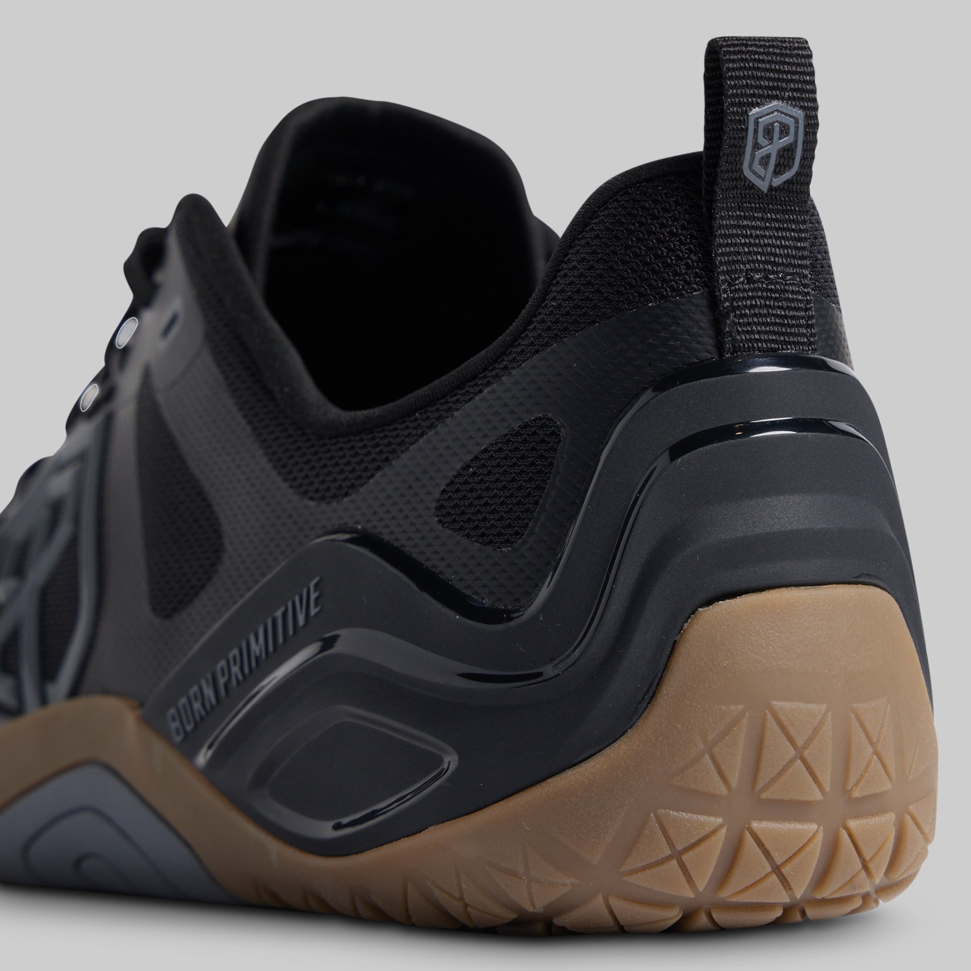 Close-up rear three-quarter view of a black Born Primitive athletic shoe with a tan outsole, showing the heel, pull tab, and textured grip pattern.