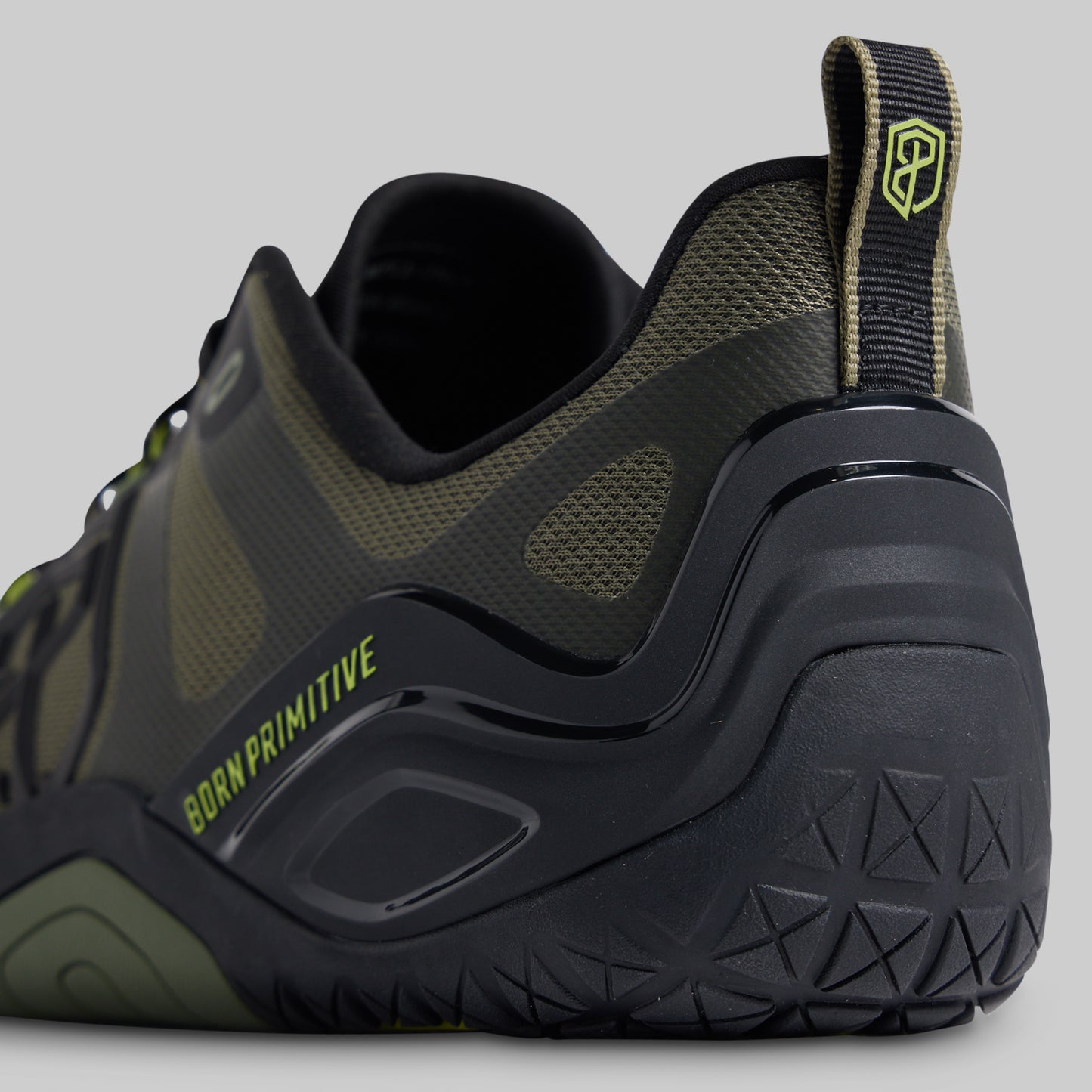 A close-up side view of a rugged athletic shoe in black and olive green, featuring a textured black sole, mesh panels, and lime green branding text that reads 'BORN PRIMITIVE' and 'P' logo on the heel pull tab.