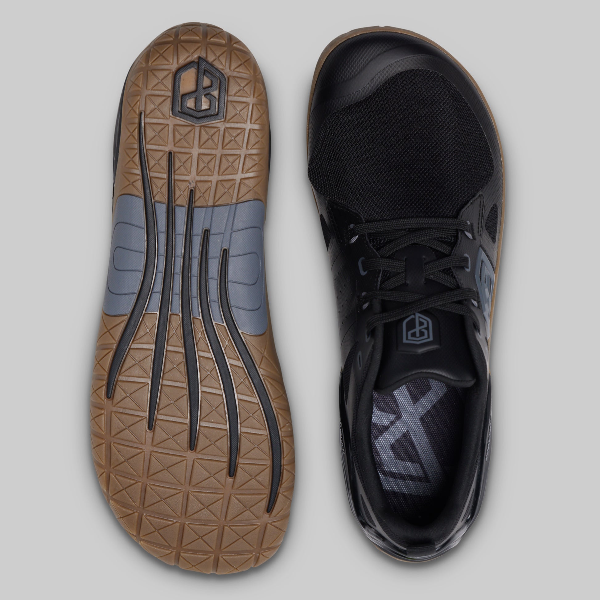 A pair of black minimalist athletic shoes shown from the top and bottom against a light gray background. The left shoe displays a brown textured outsole with gray accents, and the right shoe displays its black upper with a matching gray logo on the tongue.