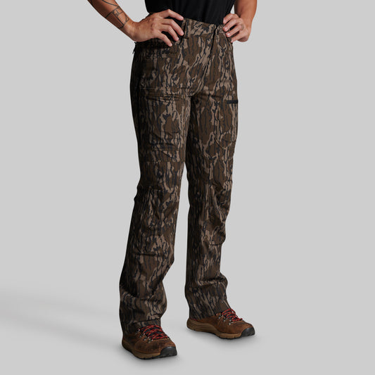 A person wearing camouflage hunting pants and brown hiking boots, standing against a light gray studio background.