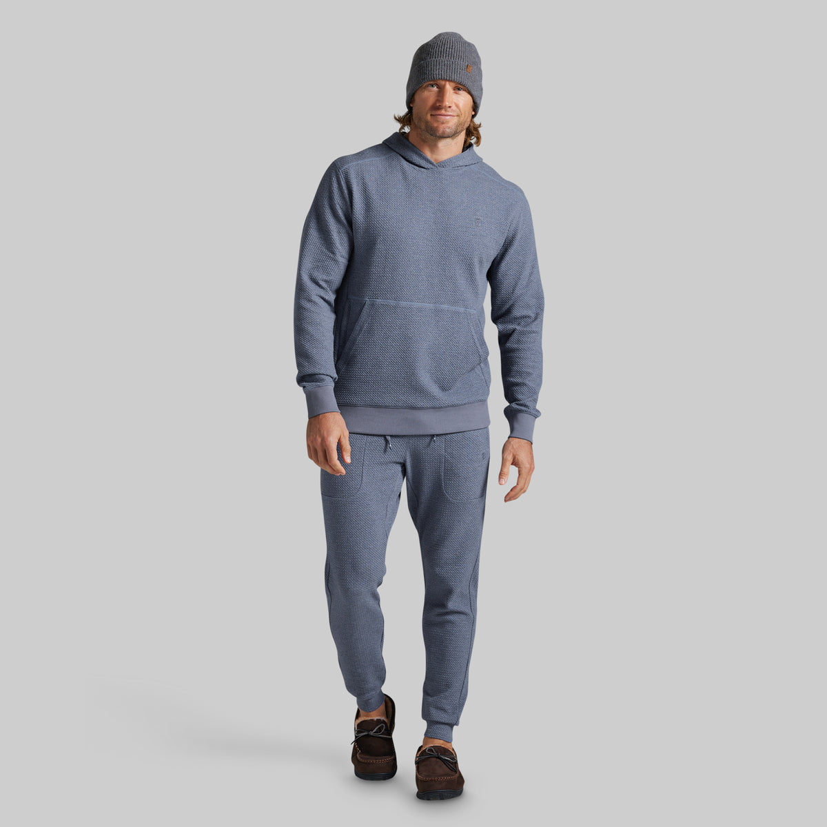 A man modeling a matching heathered slate blue hoodie and jogger sweatpants set, paired with a gray beanie and brown shoes, against a solid gray background.