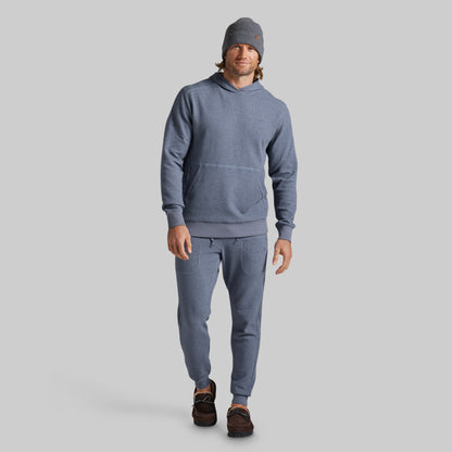 A man modeling a matching heathered slate blue hoodie and jogger sweatpants set, paired with a gray beanie and brown shoes, against a solid gray background.