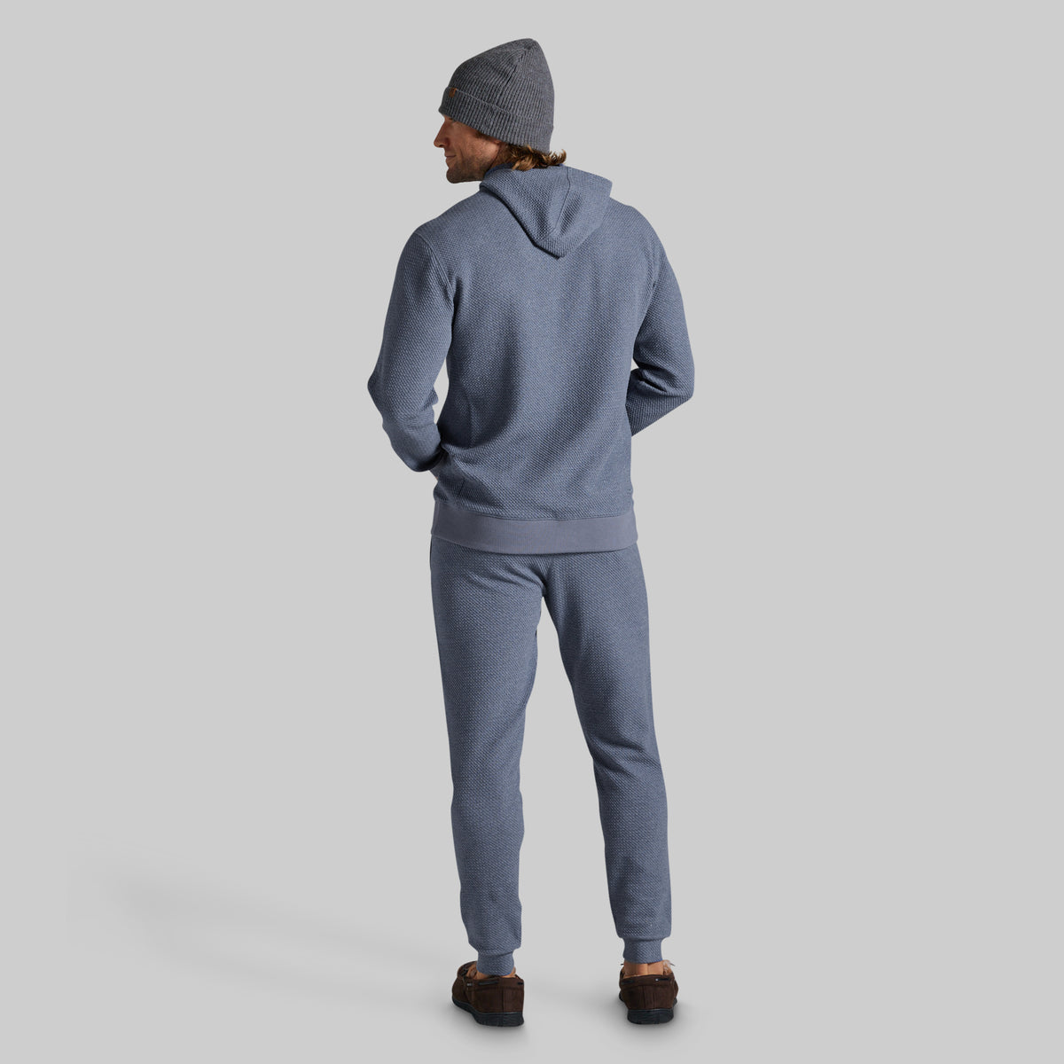 A man wearing a matching heathered slate blue hoodie and sweatpants set, viewed from behind, wearing a gray beanie and brown shoes, against a solid light gray background.