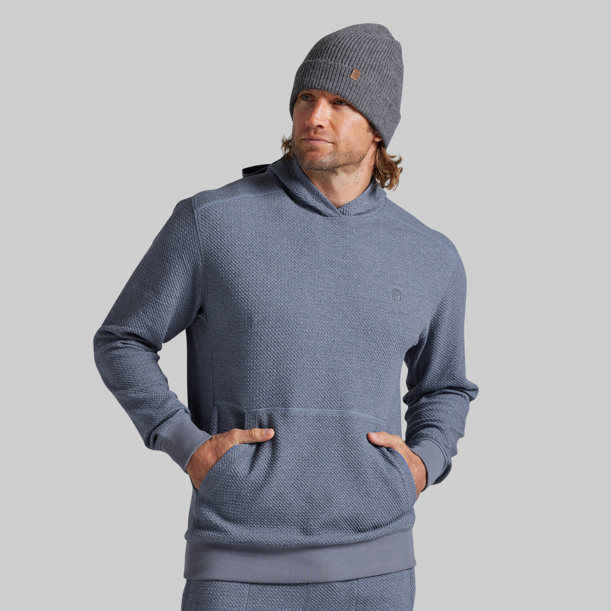 A man wearing a textured slate blue hooded sweatshirt and gray beanie against a solid gray background.