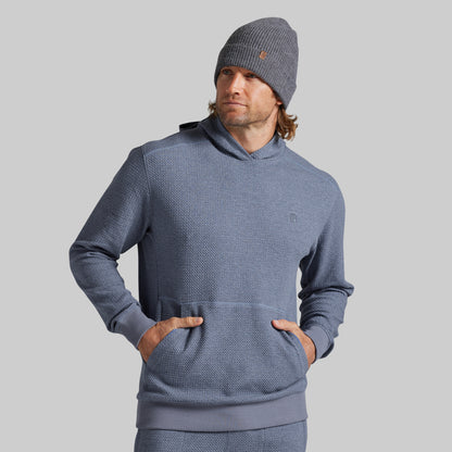 A man wearing a textured slate blue hooded sweatshirt and gray beanie against a solid gray background.
