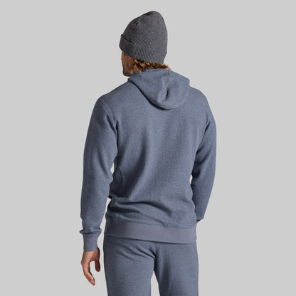 A man wearing a heathered slate blue hoodie and matching sweatpants, viewed from behind, wearing a gray knit beanie against a solid light gray background.