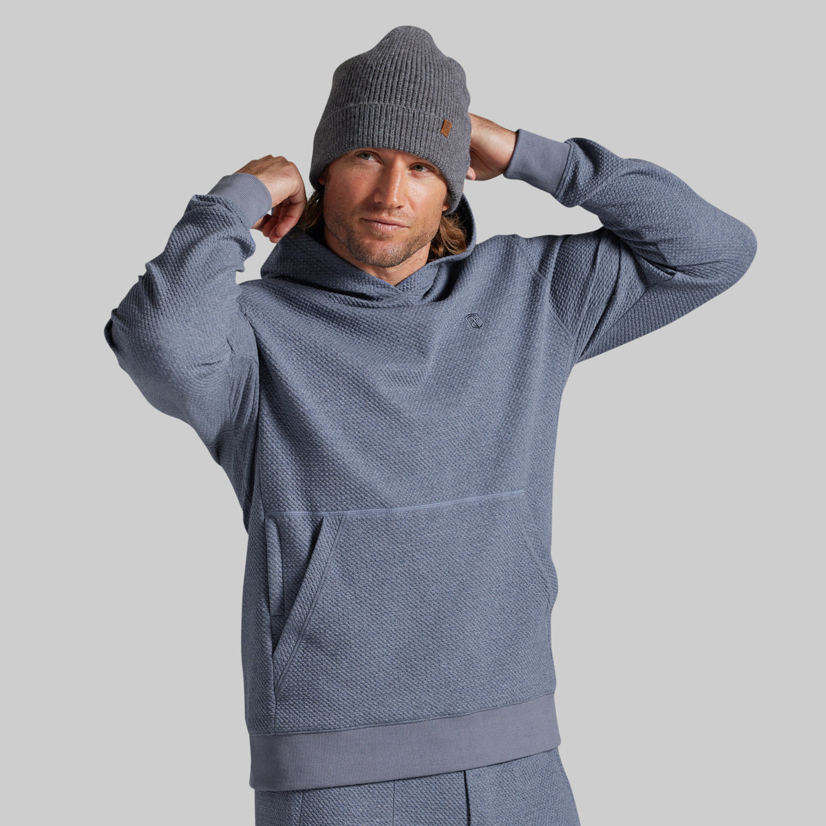A man wearing a textured slate blue hooded sweatshirt and gray beanie, posing with his hands behind his head against a solid gray background.