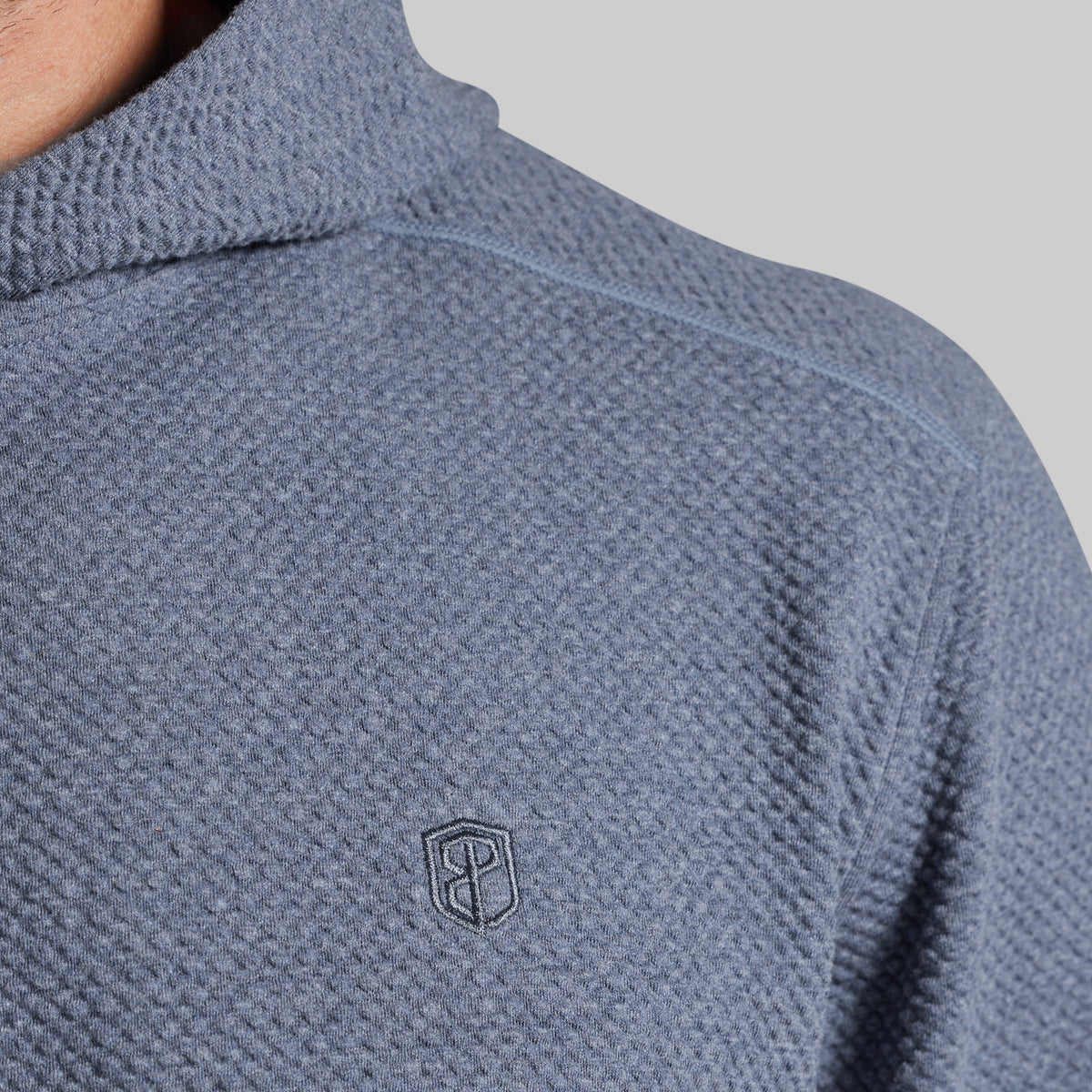 Close-up of a textured waffle-knit pullover top in a muted slate blue-gray color, featuring a small dark embroidered shield logo on the chest.